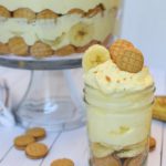 Nutter Butter No-Bake Banana Pudding inside of a small mason jar topped with miniature nutter butter cookies and trifle dish behind jar of banana pudding