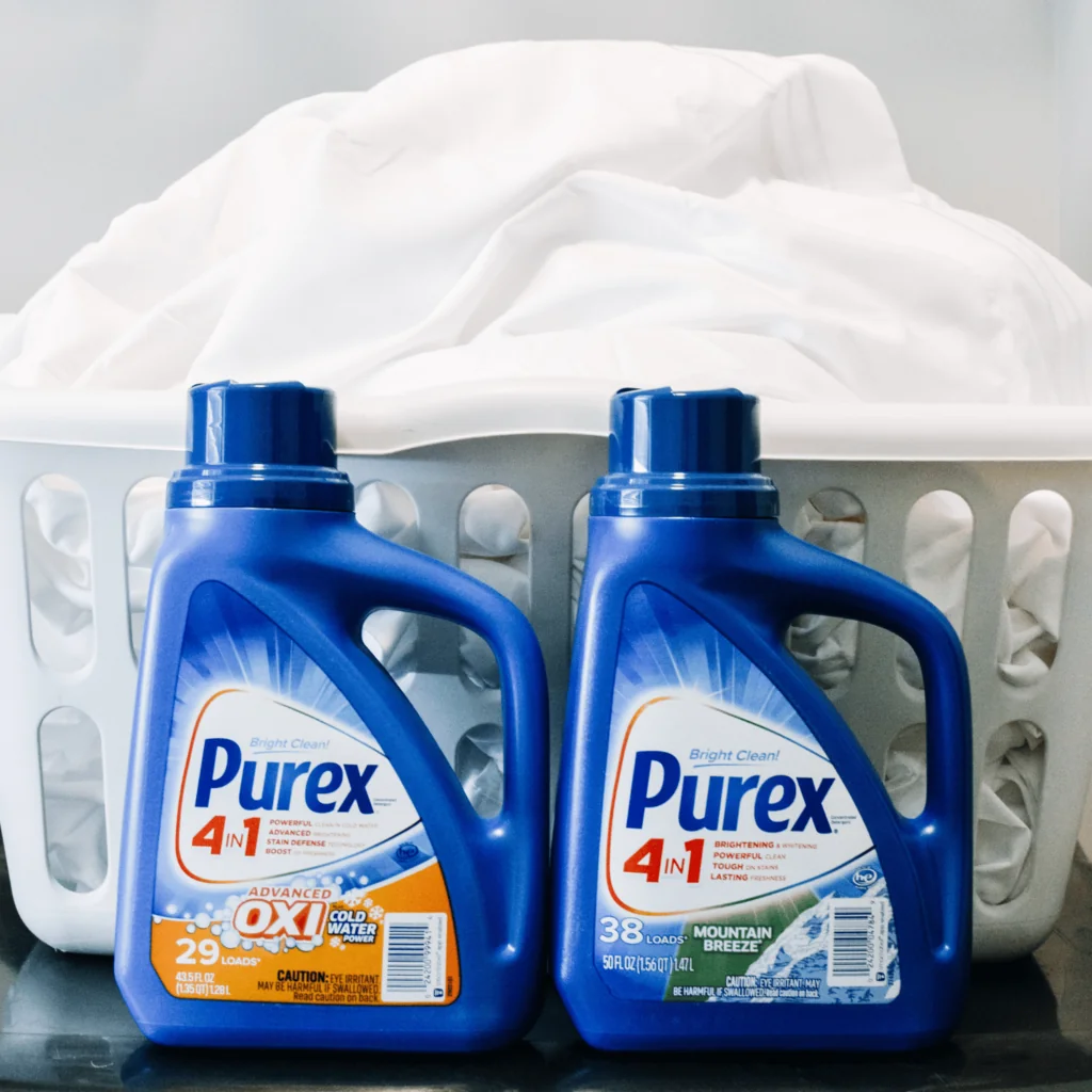 Two bottles of Purex laundry detergent next to laundry basket of white sheets for ideas on how to save on laundry costs. 
