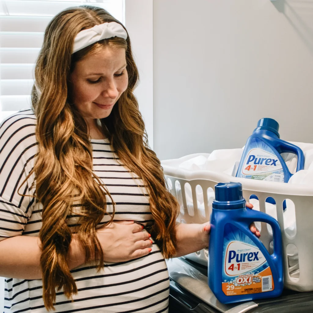 Pregnant woman next to laundry detergent and basket of sheets. 